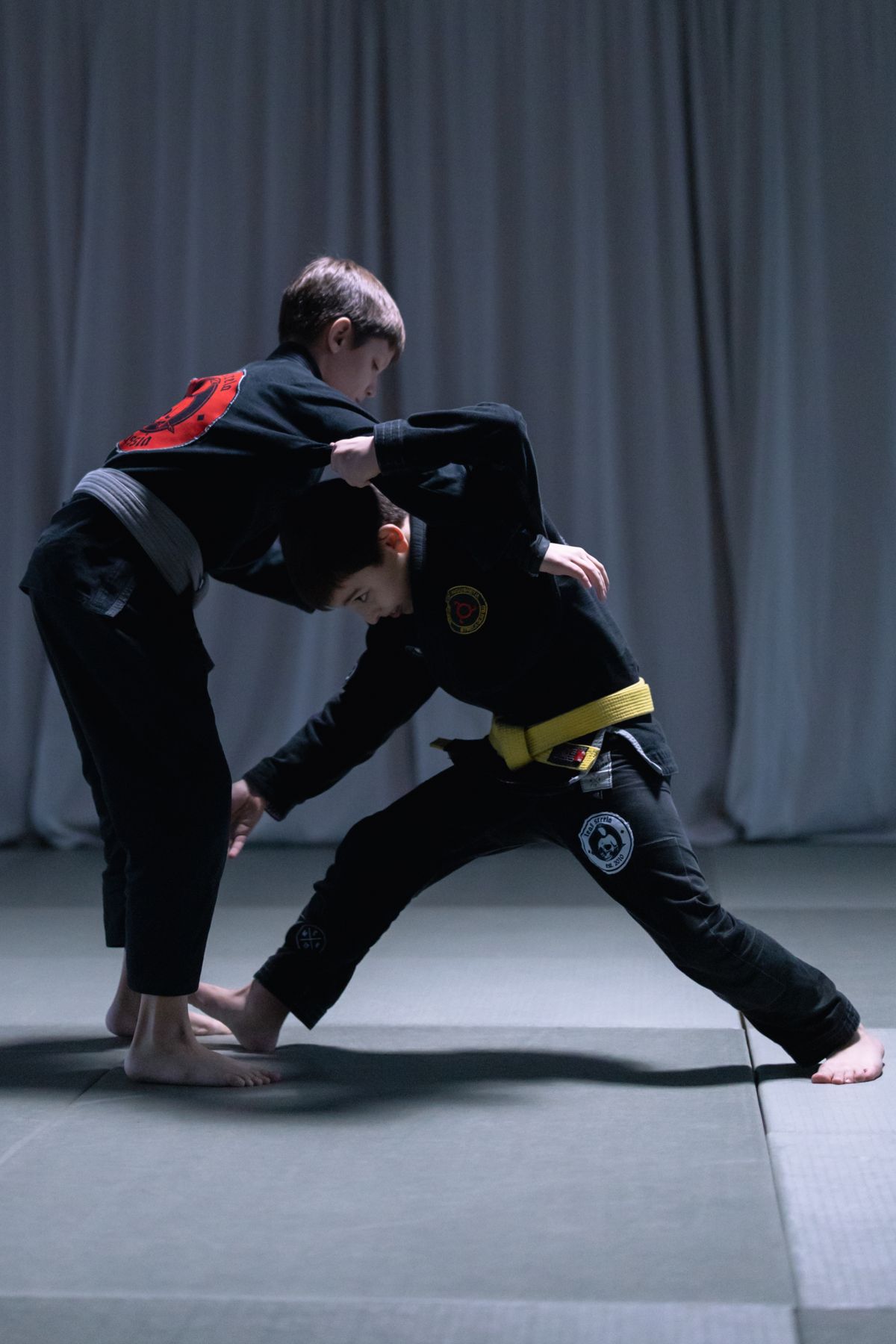 Two kids in dark gis drilling a controlled grappling technique on the mat
