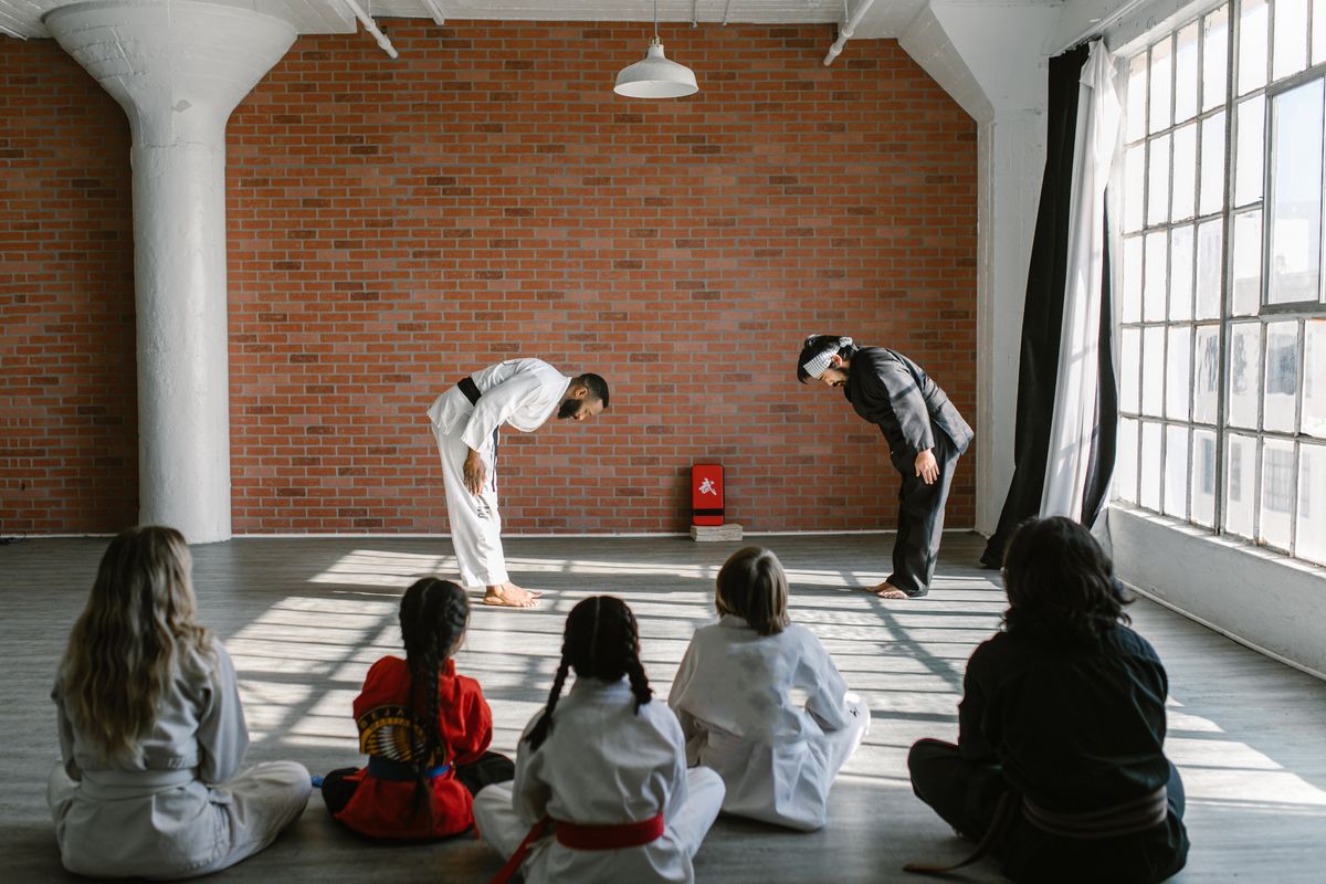 Two instructors bowing at the front of a sunlit dojo while students sit and watch
